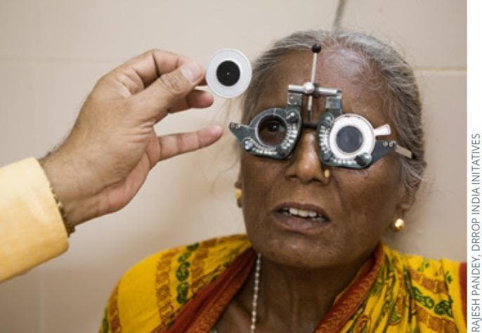 Clinical photograph of a patient undergoing a subjective refraction and visual acuity assessment. The patient is wearing an adjustable metal trial frame, an ophthalmic device featuring multiple slots designed to hold trial lenses for determining a patient's refractive error (myopia, hyperopia, or astigmatism). The examiner's hand is visible holding a pinhole occluder toward the patient's right eye. This specific tool is used in clinical practice to determine if a reduction in visual acuity is due to a refractive error—which typically improves when viewing through a small aperture—or an underlying organic pathology. The image demonstrates key steps in an optometric or ophthalmological examination, focusing on the diagnostic equipment used for determining the best-corrected visual acuity (BCVA). The setting is an eye care clinic, highlighting the procedural interaction between the clinician and the patient during vision screening.