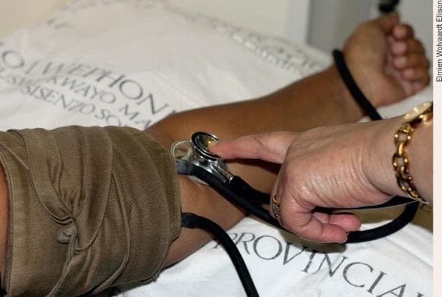 This clinical photograph demonstrates the manual assessment of blood pressure using a sphygmomanometer and an acoustic stethoscope. The image shows a healthcare provider's hand stabilizing a metal stethoscope diaphragm over the patient's antecubital fossa, specifically positioned over the brachial artery to auscultate Korotkoff sounds. A brown fabric inflatable cuff is securely wrapped around the patient's upper arm, proximal to the stethoscope placement. The patient's arm is resting on a white hospital bedsheet with printed black text. Key anatomical landmarks include the inner elbow (antecubital space) and the distal upper arm. This visual serves as an educational aid for clinical skills training, illustrating the correct placement of instruments for non-invasive blood pressure (NIBP) measurement. The medical specialty is general medicine or nursing, focusing on basic physiological monitoring and clinical examination techniques.