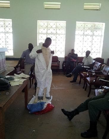 This clinical training photograph depicts a healthcare worker demonstrating the donning or doffing procedure of Personal Protective Equipment (PPE) during an Ebola Virus Disease (EVD) preparedness workshop. The central figure is shown wearing a white, full-body fluid-resistant coverall (Tyvek suit) with a front zipper closure. The participant is positioned on a designated 'clean' mat or bag to prevent environmental contamination, with tall rubber boots placed nearby for the next step of the protocol. The training takes place in an indoor classroom setting where other health professionals or immigration staff are seated in the background, observing and taking notes. A side table holds additional training materials and binders. This visual illustrates standard infection prevention and control (IPC) measures, specifically barrier nursing techniques and PPE protocols essential for managing viral hemorrhagic fevers (VHFs) in high-risk border regions.