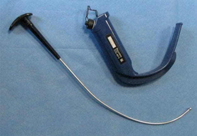 This clinical photograph displays a GlideScope video laryngoscope (GVL) alongside its dedicated rigid stylet, placed against a blue sterile field. The GVL is a reusable anesthetic device featuring an integrated, dark blue ergonomic handle and a blade with a characteristic 60-degree angulation. This significant curvature is designed to provide an indirect view of the glottis without requiring the alignment of oral, pharyngeal, and laryngeal axes. A white identification label is visible on the handle, and a connection port for a video monitor cable is located at the proximal end. Adjacent to the laryngoscope is the GlideScope-specific rigid stylet, which consists of a metallic rod with a specialized distal curve and a rounded tip to facilitate endotracheal tube placement. The stylet features a black T-shaped proximal handle for clinician control. This equipment is primarily used in airway management for both routine and difficult tracheal intubations to enhance laryngeal visualization while minimizing the upward lifting force required compared to traditional Macintosh direct laryngoscopy.