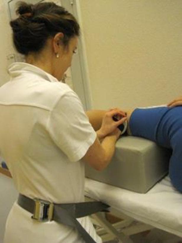This clinical photograph demonstrates a standardized procedure for measuring muscle strength using hand-held dynamometry (HHD), specifically focusing on hip extension. The patient is positioned in a side-lying posture on a medical examination table. A large, grey rectangular support block is placed between the patient’s legs to maintain neutral alignment and provide stability for the upper limb during the assessment. The examiner, wearing a white uniform, is seen applying a hand-held dynamometer to the posterior aspect of the patient's lower leg, just proximal to the ankle joint. A notable technical feature shown is the use of a wide stabilization belt worn by the examiner around their waist and anchored to the table. This belt serves as a mechanical brace to counteract the force exerted by a strong patient, ensuring that the measurement reflects the patient's actual peak force rather than being limited by the physical strength of the tester. This procedural setup is indicative of clinical research or advanced physiotherapy protocols aimed at improving the reliability and validity of objective muscle strength testing.