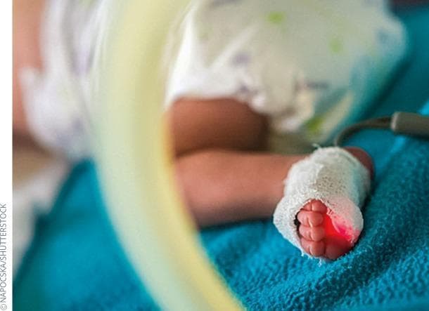 A clinical photograph depicting the neonatal intensive care of a preterm infant, focusing on the application of a pulse oximetry probe to the foot. The probe is secured across the midfoot and digits with a white, porous medical-grade adhesive wrap or gauze, which emits a characteristic red light from the internal light-emitting diode (LED) used for spectrophotometric oxygen saturation (SpO2) measurement. A thin, gray data cable extends from the probe to a bedside monitor (not visible). The infant's lower extremity is resting on a textured blue fabric surface, typical of neonatal bedding, with a translucent yellow medical tube visible in the blurred foreground, likely part of an incubator or respiratory support system. This image demonstrates standard non-invasive hemodynamic monitoring in a pediatric or neonatal clinical setting, emphasizing the importance of continuous oxygenation assessment in preterm newborns.