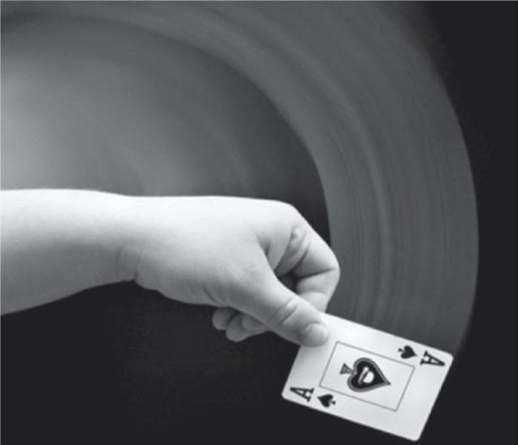 This grayscale educational photograph demonstrates the 'motion streak' phenomenon, a key concept in visual psychophysics and neurobiology. The image features a human hand holding an Ace of Spades playing card, captured while moving in a rapid arcing motion. A distinct blurred trail, or motion streak, extends behind the path of the card, creating a curved, translucent visual index of the object's trajectory. This visual is used to illustrate form-motion integration within the human visual system, specifically the interaction between the ventral stream (the 'what' pathway, responsible for identifying the card's suit and rank) and the dorsal stream (the 'where/how' pathway, responsible for processing motion and spatial orientation). The phenomenon arises from retinal persistence, where the brain utilizes oriented blur to assist in the perception of fast-moving stimuli. This illustration is significant for medical and psychological studies focusing on visual perception, neurophysiology of the striate and extrastriate cortex, and the functional organization of parallel visual processing streams.