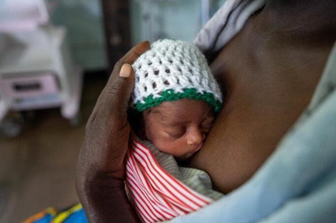 This clinical photograph demonstrates the application of Kangaroo Mother Care (KMC) for a low-birth-weight neonate in a hospital setting. The image shows a preterm infant held in direct skin-to-skin contact against the mother's chest, a critical intervention for thermal regulation, bonding, and physiological stability. The neonate is wearing a white crocheted cap with a green trim to prevent heat loss through the head. The infant is secured in an upright position using a red-and-white striped cloth wrap, with the mother's hand providing additional support to the head. This visual illustrates key components of small and sick newborn care (SSNC), specifically addressing hypothermia prevention and essential newborn care in resource-limited environments. The background includes a neonatal cot, indicating the clinical environment of a Neonatal Intensive Care Unit (NICU). This image serves as an educational example of non-pharmacological interventions used to improve survival outcomes in premature or low-birth-weight infants.