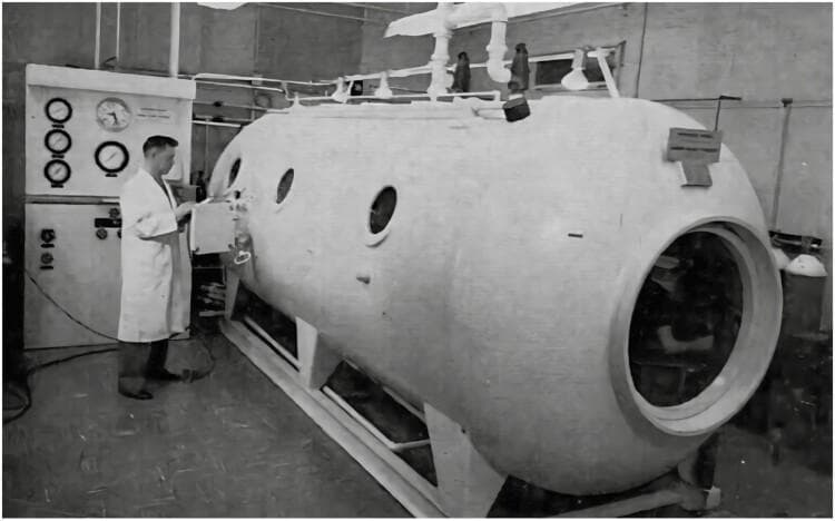 A clinical photograph of a multi-place hyperbaric oxygen therapy (HBOT) or diving chamber, used for treating decompression sickness ('the bends') and conducting respiratory research. The image depicts a large, cylindrical pressure vessel with rounded ends, featuring multiple circular observation portholes and an entry hatch. It is mounted on a heavy-duty metal support frame. Adjacent to the chamber is a vertical control console operated by a clinician. The console is equipped with large analog pressure gauges, flow meters, and valves used to monitor and regulate simulated depth, atmospheric pressure, and gas mixtures within the chamber. The setup illustrates the technology required for hyperbaric medicine and physiological testing at high ambient pressures. Historically located at the Royal Victoria Hospital (Royal Vic), this equipment represents an essential tool in respiratory physiology and the clinical management of gas embolism and nitrogen narcosis.