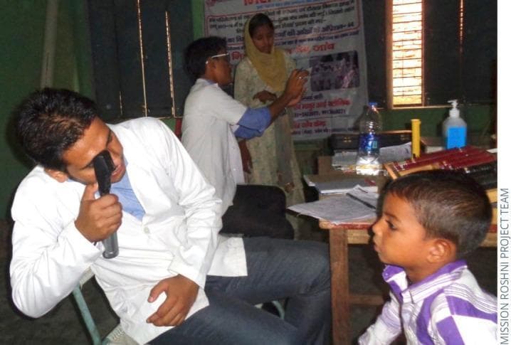 A clinical photograph depicting a pediatric ophthalmic screening in a community outreach setting. The primary focus is a male healthcare provider, wearing a white lab coat, performing a direct ophthalmoscopy on a young male patient. The clinician is using a handheld direct ophthalmoscope to visualize the internal structures of the boy's eye, including the retina, optic disc, and macula, to screen for refractive errors or ocular pathologies. The background reveals a makeshift clinic environment featuring a table with medical documentation, a case containing trial lenses for vision correction, and hand sanitizer. Additional staff are visible in the background, including one individual performing a visual acuity or near-point test. This image illustrates concepts of public health ophthalmology, community-based vision screening, and the application of diagnostic medical devices in primary care or resource-limited settings.