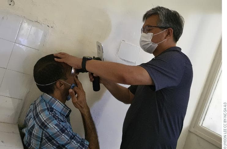 This clinical photograph demonstrates a portable ophthalmic examination being performed in a community or clinical setting. A healthcare provider is seen using a handheld digital fundus camera, integrated with a smartphone or similar imaging interface, to capture a retinal image from a seated patient. The provider, wearing a protective surgical mask and corrective lenses, stabilizes the patient's head with their left hand while positioning the device close to the patient's right eye. An active light source from the device is visible, indicating the illumination of the posterior segment for screening purposes. This visual illustrates modern diagnostic technology used for mobile screening of ocular diseases, such as diabetic retinopathy, where traditional table-mounted fundus cameras may not be accessible. The image highlights the intersection of ophthalmology, portable medical devices, and artificial intelligence-capable hardware in public health screenings.