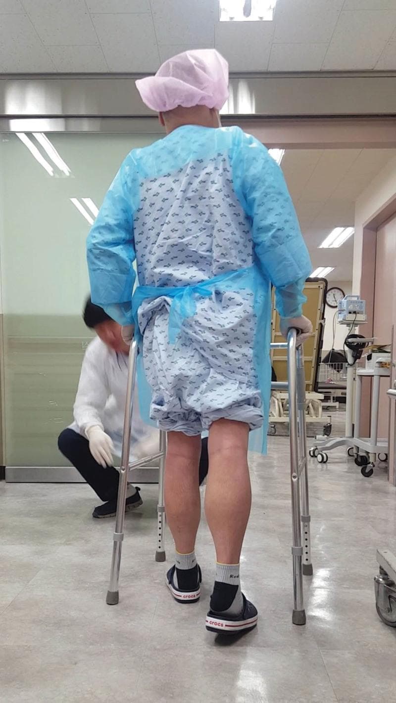 A clinical photograph depicting a postoperative patient engaged in a walker-assisted gait training session as part of a phase I cardiac rehabilitation program. The patient, shown from a rear perspective, is utilizing a standard height-adjustable silver metal walker with non-slip rubber tips. The patient is wearing protective medical attire, including a pink surgical cap, a patterned hospital gown, and a blue protective isolation gown, indicating adherence to infection control protocols following a heart transplant and midline sternotomy. The lower extremities are visible, demonstrating weight-bearing ambulation on postoperative day 28. A physical therapist or healthcare provider is seen in the background in a crouched position, providing supervision and ensuring patient safety during the mobility assessment. The clinical setting is a hospital general ward or rehabilitation area, containing medical equipment such as a tilt table and monitoring stands. This visual documents a milestone in the functional recovery and physical therapy progression after complex thoracic surgery.