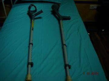 This clinical photograph displays a pair of adjustable elbow crutches, also known as Loftstrand or forearm crutches, resting on a flat teal surface. Each mobility aid consists of a contoured, semi-circular forearm cuff and an ergonomic hand grip at the top, typically made of high-density plastic or rubber. The main support structure is a metallic telescopic shaft featuring a series of adjustment holes and spring-loaded locking pins to customize the height for the user's specific anthropometrics. At the distal end, each crutch is fitted with a wide, non-slip rubber tip (ferrule) to provide traction and stability during ambulation. These devices are commonly utilized in physical therapy and orthopedic rehabilitation for patients with paraplegia, spinal cord injuries, or lower limb impairments who possess sufficient upper body strength and coordination to transition from more stable aids like walkers to less restrictive mobility tools.