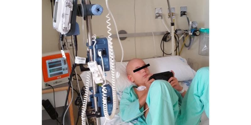A clinical photograph of a pediatric oncology patient in a hospital bed, illustrating the integration of supportive technology and distraction therapy. The patient, appearing bald (suggestive of chemotherapy-induced alopecia), is positioned in a semi-recumbent posture wearing a green gown. The primary focus is the medical setup including a Patient-Controlled Analgesia (PCA) morphine pump mounted on an IV pole and a multi-parameter monitor displaying respiratory and cardiac waveforms. Clinical connections are visible via intravenous tubing and a chest-mounted sensor for monitoring heart rate variability (HRV) and analgesia nociception index (ANI). The photograph demonstrates the use of an electronic video game (EVG) as a non-pharmacological intervention for managing incidental and basal pain related to chemotherapy-induced mucositis. The setting includes standard intensive or acute care infrastructure such as wall-mounted oxygen flowmeters and suction canisters, highlighting a multidisciplinary approach to pediatric pain management and autonomic nervous system monitoring.