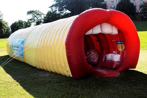 This photograph depicts a large-scale, walk-through educational tool known as a 'giant larynx' model, used in public health campaigns to teach human anatomy and voice production. The structure is an elongated inflatable tunnel with a segmented yellow exterior. One end is designed as a large open mouth featuring oversized red lips and white anatomical representations of upper teeth. The red interior visible through the mouth opening represents the pharynx and larynx, including structural interpretations of the tongue and vocal cords. This interactive anatomical diagram serves as a public education device for the specialty of otolaryngology (ENT), focusing on the upper respiratory tract and phonation. It is designed to simplify complex internal structures for a general audience, demonstrating the pathway from the mouth through the throat.