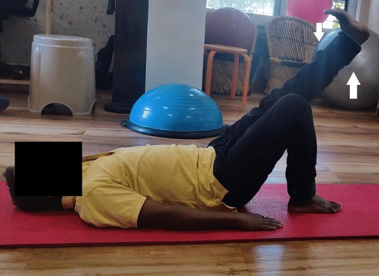 A clinical photograph depicting a male patient in a physiotherapy setting, performing a therapeutic exercise. The patient is positioned supine on a red yoga mat on a wood-laminate floor. The patient is executing a straight leg raise (SLR), with the right lower extremity actively elevated while maintaining knee extension. The contralateral (left) leg is positioned in a crook-lying posture with the knee flexed and the foot flat on the mat to stabilize the lumbar spine and pelvis. The background contains common rehabilitation equipment, including a blue Bosu balance trainer, a gray stability ball, and various stools. This visual demonstrates a post-operative or conservative management exercise focused on strengthening the hip flexors and quadriceps, frequently utilized in rehabilitation protocols for conditions such as avascular necrosis of the femoral head or following total hip replacement.