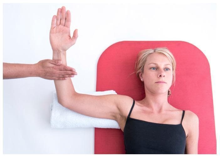 A clinical photograph illustrating a musculoskeletal physical examination technique for the shoulder. The patient is positioned supine on a red mat with the right shoulder abducted to 90 degrees and the elbow flexed to 90 degrees. A rolled white towel is placed under the distal humerus to stabilize the joint and maintain neutral alignment. An examiner's hand is placed on the anterior aspect of the patient's distal forearm, applying resistance as the patient attempts internal rotation. This maneuver is used to assess the strength and integrity of the anterior rotator cuff muscles, specifically the subscapularis, and to identify potential pathology such as tendinopathy or muscle tears. The image demonstrates proper patient positioning and hand placement for orthopedic testing in a physical therapy or clinical setting.