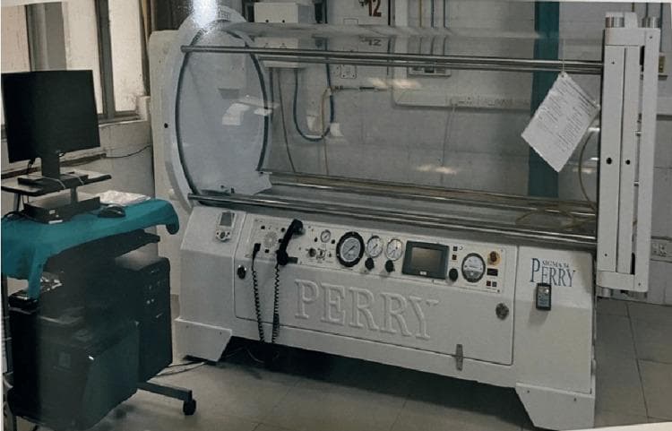 This clinical photograph displays a monoplace hyperbaric oxygen therapy (HBOT) chamber, a specialized medical device used to deliver 100% oxygen at pressures greater than sea level. The unit features a large, transparent acrylic cylindrical pressure hull, allowing for continuous visual monitoring of a single patient. The chamber's base is a white-enameled metal frame (branded 'PERRY') equipped with a comprehensive integrated control console. This panel includes analog pressure gauges, a digital display screen, manual control valves, and a communication handset for patient-operator interaction. To the left, a mobile computer workstation is positioned for data logging and session monitoring. This equipment is primarily utilized in wound care, emergency medicine, and hyperbaric medicine departments for treating conditions such as chronic non-healing ulcers (e.g., diabetic foot ulcers), decompression sickness, and carbon monoxide poisoning. The setup illustrates the standard clinical environment for administering adjunctive hyperbaric therapy.