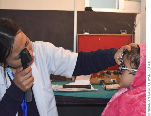 A clinical photograph illustrating a pediatric ophthalmic examination, specifically demonstrating the technique for 'wet' cycloplegic retinoscopy. A female medical professional in a white coat is shown in the left of the frame, holding a handheld retinoscope to her right eye. She is aiming the light through a trial frame fitted onto a young child positioned on the right. The practitioner uses her left hand to stabilize the trial frame on the child's face, ensuring proper alignment for refraction. The child is wearing a pink hooded garment. This procedure is typically performed after the administration of cycloplegic agents, such as cyclopentolate or tropicamide, to paralyze the ciliary muscle and prevent active accommodation, thereby allowing for an accurate objective measurement of the patient's refractive error. The background shows clinical supplies, including a lens trial set, indicating a professional optometric or ophthalmic setting.