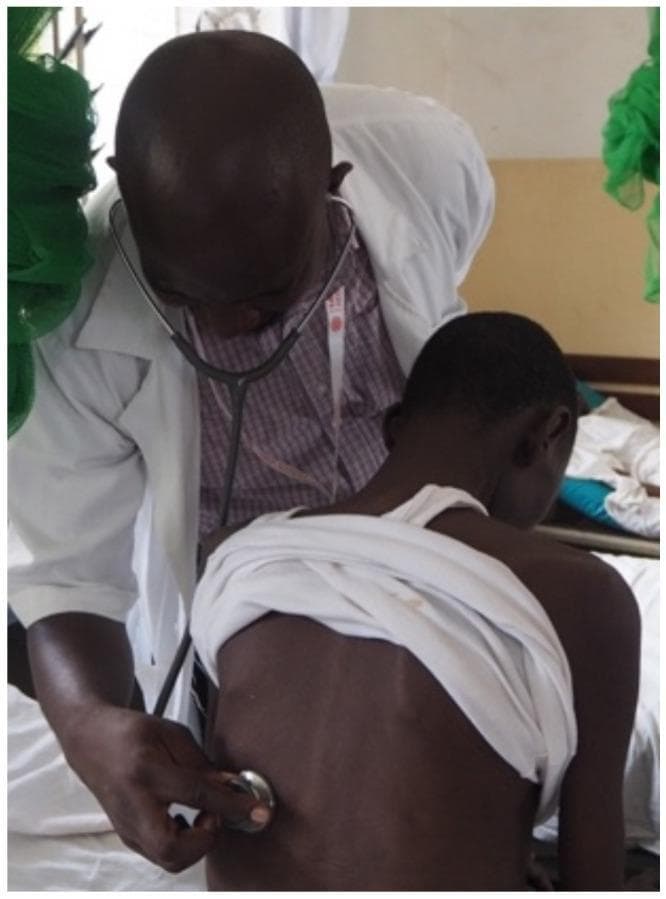 This clinical photograph captures a healthcare professional performing a respiratory physical examination on a patient in a ward setting. The physician, wearing a white clinical coat and patterned shirt, is seen leaning forward in a focused posture. He is using an acoustic stethoscope to perform auscultation of the patient's posterior chest (back). The patient is seated on a hospital bed, facing away from the camera, with a white cloth draped around their shoulders to expose the thoracic region for examination. The image illustrates standard clinical practice for evaluating pulmonary health, particularly relevant in the context of screening for opportunistic infections like tuberculosis in HIV-positive populations within resource-limited settings. The setting appears to be an inpatient or outpatient department specialized in infectious disease management.