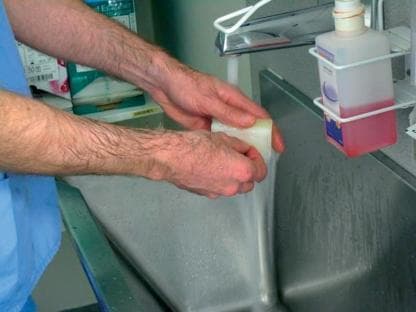 A clinical photograph demonstrating a fundamental infection control procedure: surgical hand antisepsis or clinical handwashing. The image shows a healthcare professional wearing blue scrubs, with arms exposed to the mid-forearm, performing hand hygiene at a stainless steel medical scrub sink. A steady stream of water flows from a chrome faucet. The individual is actively lathering a white rectangular surgical scrub sponge or bar of soap between their hands. To the right of the faucet, a wall-mounted wire rack holds a plastic dispenser bottle containing a reddish-pink antimicrobial solution, likely chlorhexidine gluconate or povidone-iodine. The background shows clinical supply packaging, reinforcing the hospital or surgical setting. This visual illustrates standard precautions, aseptic technique, and the prevention of nosocomial infections in a clinical environment.