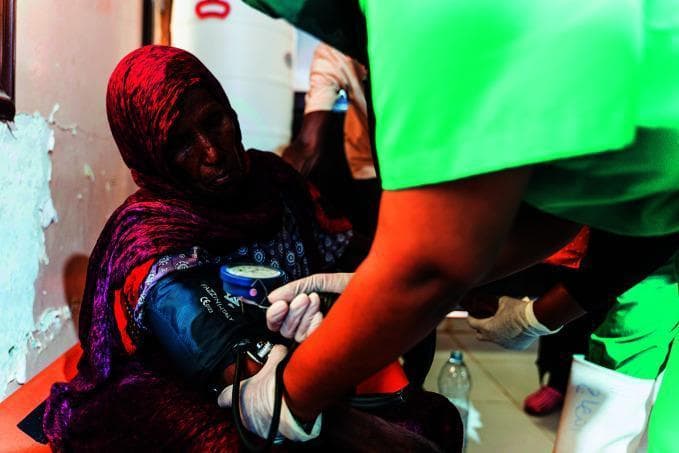 This clinical photograph captures a healthcare interaction in a field medical setting, specifically a cholera treatment center. The image shows a healthcare provider performing a non-invasive blood pressure measurement on a seated adult female patient. The clinician, wearing nitrile gloves and green scrubs, has positioned an aneroid sphygmomanometer cuff on the patient's left upper arm. A visible blue-faced manual pressure gauge is attached to the cuff. The provider is simultaneously using a stethoscope (partially visible) for auscultation of Korotkoff sounds to determine systolic and diastolic values. This scene illustrates vital sign monitoring as a critical component of clinical assessment and hemodynamic stabilization in infectious disease management, particularly for monitoring dehydration levels in cholera patients. The setting emphasizes healthcare delivery in resource-limited or humanitarian crisis environments.