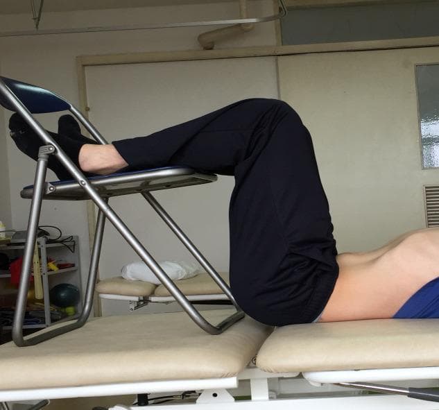 This clinical photograph demonstrates a participant performing an abdominal hollowing exercise, a therapeutic technique used in physical therapy and sports medicine to activate deep core stabilizers like the transversus abdominis. The subject is positioned supine on a padded examination table in a '90-90' posture. This involves 90 degrees of hip flexion and 90 degrees of knee flexion, with the lower legs and feet supported on the seat of a folding chair to neutralize the lumbar spine. The image captures the visible depression or 'hollowing' of the lower abdominal wall toward the spine, contrasting with the relatively stable position of the upper torso and limbs. This visual demonstrates the clinical application of core muscle recruitment protocols often used in rehabilitative research for low back pain management. The setting appears to be a clinical or research laboratory environment, emphasizing controlled positioning for consistent muscle activation measurements.