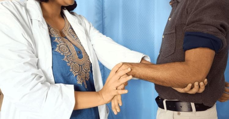A clinical photograph illustrating the performance of Mill’s test, a provocative physical examination maneuver used to diagnose lateral epicondylitis (tennis elbow). The image shows a healthcare professional in a white coat interacting with a male patient. The clinician is standing, using one hand to palpate the patient's lateral epicondyle at the elbow, while the other hand grasps the patient's hand to maintain the wrist in a flexed position with the forearm pronated. The patient's arm is extended at the elbow. This specific positioning—passive pronation of the forearm, flexion of the wrist, and extension of the elbow—is designed to stretch the extensor carpi radialis brevis muscle, which elicits pain in positive cases of lateral epicondylitis. The educational focus is on orthopedic assessment techniques and musculoskeletal physical examination. The background features a clinical setting with a blue curtain.
