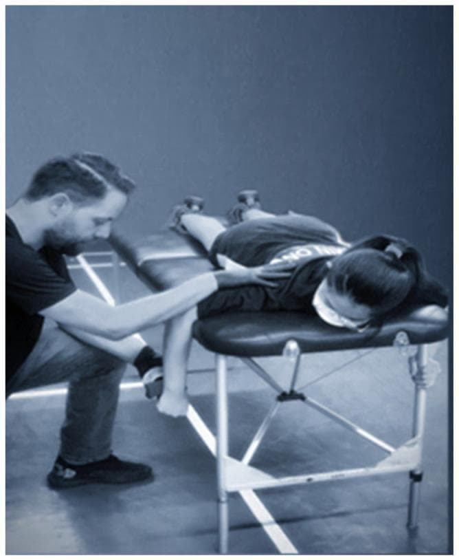 Clinical photograph demonstrating a shoulder internal rotation strength assessment using a Hand-Held Dynamometer (HHD) in a physical therapy or sports medicine setting. The subject is positioned prone on a padded examination table with the head turned away from the testing side. The humerus of the tested arm is abducted to 90 degrees and supported by the edge of the table, while the elbow is flexed at 90 degrees, allowing the forearm to hang vertically. An examiner is shown kneeling beside the patient, stabilizing the subject's scapula/humerus with one hand to prevent compensatory movements, while applying the HHD to the anterior surface of the distal forearm, proximal to the ulnar styloid process, with the other hand. This setup is specifically designed to isolate and measure the isometric strength of the internal rotator muscle group (primarily the subscapularis, pectoralis major, and latissimus dorsi) for clinical evaluation of muscle imbalances or rehabilitation progress in athletes.