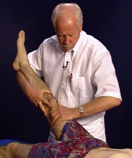 A clinical photograph demonstrating the performance of the Lasègue test, also known as the straight leg raise test. The image depicts a male examiner in a white short-sleeved shirt performing a passive physical examination on a supine patient. The examiner is positioned at the patient's side, using both hands to support and elevate the patient's right lower extremity while maintaining knee extension. One hand is placed on the distal thigh/suprapatellar region, and the other provides support under the calf/achilles area to facilitate gradual hip flexion. The patient's foot is in a neutral or slightly dorsiflexed position. This orthopedic and neurological assessment is clinically used to evaluate for lumbar radiculopathy, specifically irritation of the sciatic nerve or nerve roots (L4-S1), which may be caused by conditions such as intervertebral disc herniation. The test is considered positive if radicular pain is reproduced in the leg between 30 and 70 degrees of hip flexion. The educational focus is on the correct manual technique and patient positioning for identifying signs of nerve root compression or meningeal irritation.