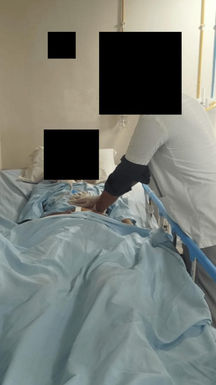 This clinical photograph illustrates a healthcare professional performing chest physiotherapy on a patient in a hospital setting. The patient is positioned supine in a hospital bed, with the clinician standing at the bedside applying a manual chest vibration technique. The clinician's hands are placed specifically over the patient's left lower lung zone, a maneuver intended to facilitate the mobilization and clearance of pulmonary secretions. The patient appears to be receiving supportive care, evidenced by the presence of medical tubing (likely an endotracheal or nasogastric tube) and hospital attire. The clinician is shown wearing protective gloves while performing the intervention. This image serves as an educational example of active pulmonary rehabilitation and airway clearance therapy within a clinical or post-operative context, highlighting manual techniques used in respiratory physiotherapy.