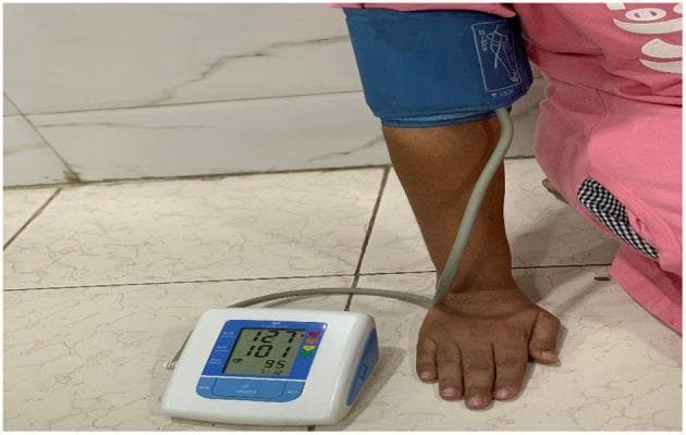 A clinical photograph demonstrating the measurement of vital signs using an automated digital sphygmomanometer on a human patient. The setup includes a blue inflatable compression cuff wrapped around the patient's upper arm, connected via flexible gray tubing to a portable digital monitor. The monitor features a liquid crystal display (LCD) showing three stacked numerical values: 127 (systolic pressure), 101 (diastolic pressure), and 95 (pulse rate), accompanied by colored heart icons and time indicators. This device is used for non-invasive blood pressure (NIBP) monitoring and pulse rate assessment, essential in evaluating cardiovascular health and risk factors such as hypertension. The patient is positioned with their arm resting on a flat surface to ensure accurate measurement levels. This visual represents standard clinical practice for gathering biometric data in both primary care and remote health monitoring settings.