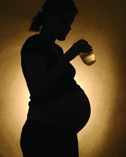 A clinical educational photograph utilizing a silhouette technique to represent a pregnant woman in her second or third trimester. The individual is shown in profile, with a visibly enlarged abdomen indicating advanced pregnancy. She is holding a glass of liquid to her mouth, symbolizing the ingestion of water or other substances. From a medical and public health perspective, this image serves as a conceptual illustration of prenatal environmental exposures. It is used to teach concepts related to maternal-fetal health, toxicology, and environmental health risks, specifically the impact of contaminated drinking water (e.g., perfluorooctanoic acid or PFOA) on gestational outcomes such as pregnancy-induced hypertension or preeclampsia. The educational focus is on the vulnerability of the maternal-fetal unit to ingested environmental pollutants and the importance of safe hydration during pregnancy.