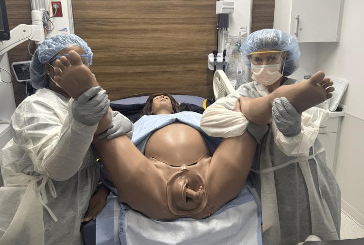 This clinical photograph displays a high-fidelity obstetric patient simulator (Victoria S2200) being used to demonstrate the McRoberts maneuver in a dorsal lithotomy position. The simulator is positioned on a delivery bed with the legs hyperflexed toward the abdomen to rotate the pelvis cephalad and flatten the lumbar spine, a technique primarily utilized to manage shoulder dystocia or facilitate breech deliveries. Two clinical staff members in full personal protective equipment (gowns, masks, caps, and face shields) are manually supporting the simulator's legs in the hyperflexed position. At the vaginal opening, the simulator demonstrates a footling breech presentation, with a single fetal foot visible emerging from the birth canal. This visual serves as an educational tool for medical training in emergency obstetrical maneuvers, highlighting the anatomical positioning necessary to optimize the pelvic outlet and the visualization of complex fetal presentations during vaginal delivery.