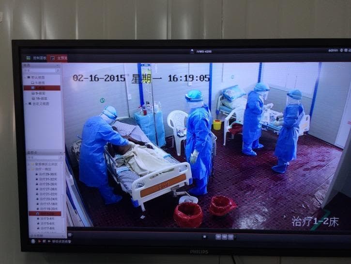 Clinical photograph of a high-containment Ebola Treatment Unit (ETU) ward, captured via a surveillance monitor. The image illustrates advanced infection control measures during the management of a highly infectious viral hemorrhagic fever. Multiple medical personnel are visible wearing full-body Level C/D personal protective equipment (PPE), consisting of fluid-resistant blue coveralls with integrated hoods, full-face shields, and waterproof shoe covers. They are shown providing direct bedside care to a patient on a clinical hospital bed. The environment is partitioned for biocontainment, featuring red-lined biohazard waste receptacles for contaminated materials and intravenous therapy equipment. This visual emphasizes the stringent protocols required for the containment of infectious diseases like Ebola, highlighting the layout of a contaminated zone and the operational requirements of healthcare workers in a specialized treatment center.