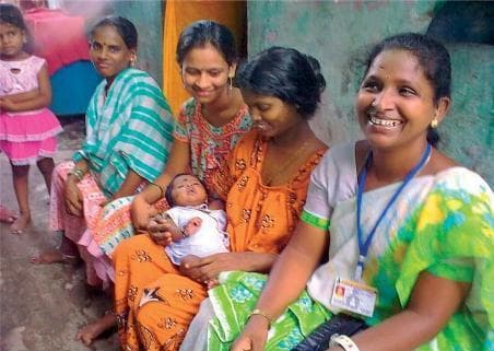 A clinical photograph depicting a community-based maternal and child healthcare outreach setting in Mumbai, India. The image shows a group of women and a young child, with a female eye care worker positioned in the foreground on the right. The healthcare worker is identified by her professional blue lanyard and identification badge, which includes a photo and text, worn over a traditional green and white patterned sari and white blouse. She is smiling, indicative of a supportive community engagement role. Adjacent to her, a mother holds an infant dressed in a white shirt; the infant is positioned supine and appears to be under observation or receiving community-level postnatal support. This image illustrates the delivery of primary health services, specifically community eye care and maternal-child health screening, by trained local health workers within residential urban environments. It highlights concepts of healthcare accessibility, community health workers (CHWs), and primary prevention in global health contexts.