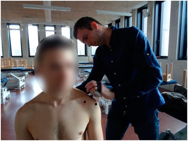 A clinical photograph depicting a physical examination of the shoulder joint in a medical or rehabilitative setting. An examiner is shown using a digital pressure algometer to quantify the pressure pain threshold (PPT) at the acromioclavicular (AC) joint of a seated patient. Three specific anatomical landmarks on the anterior and superior shoulder are indicated by black circular markers: the coracoid process, the AC joint, and the anterolateral subacromial area. The image demonstrates a standardized diagnostic procedure often used in the assessment of musculoskeletal conditions such as adhesive capsulitis (frozen shoulder) or subacromial impingement syndrome. The focus is on the correct placement of the algometer and the topographical identification of myofascial or articular trigger points for clinical evaluation of pain sensitivity and joint pathology.