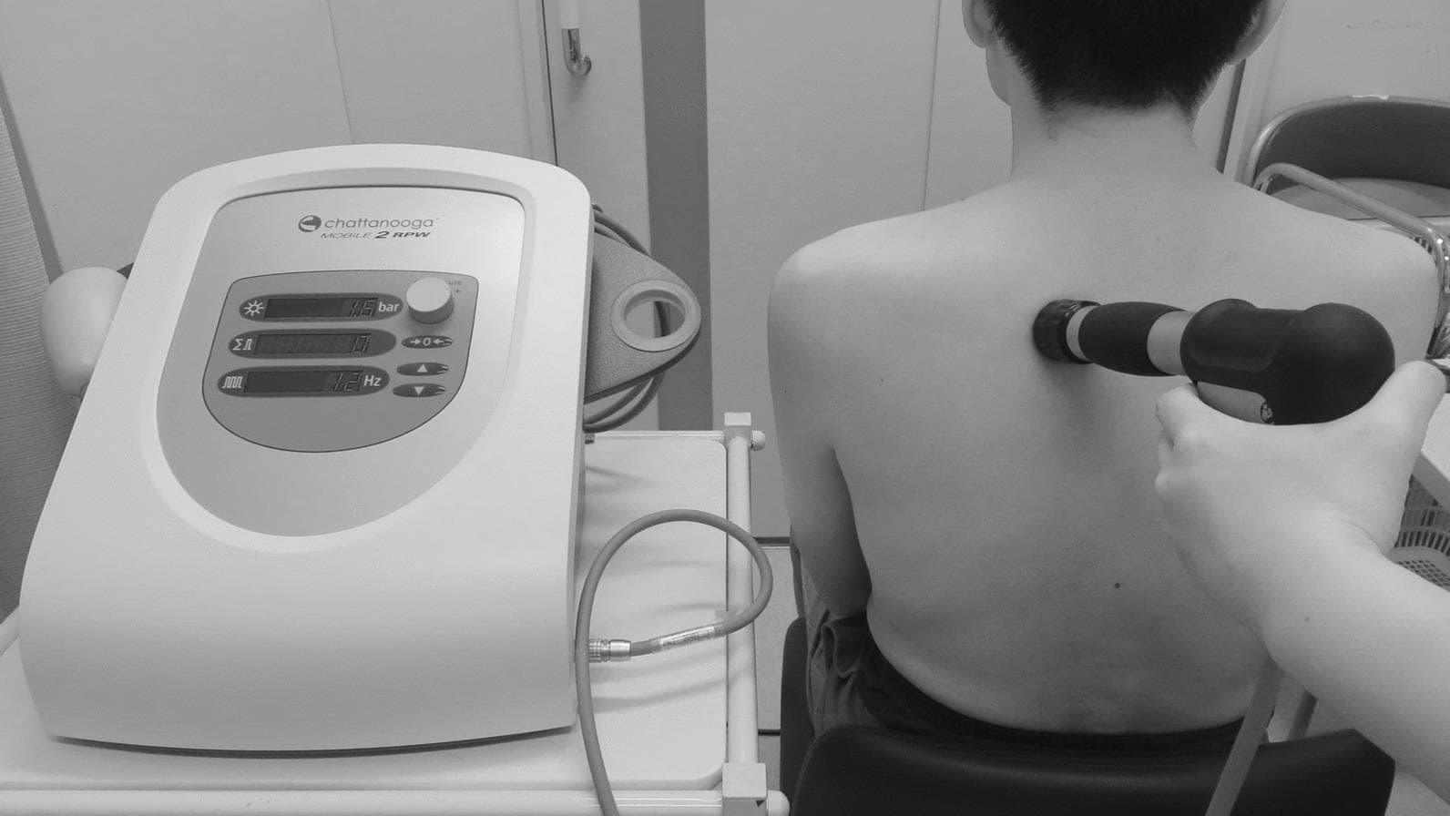 This procedural photograph illustrates the clinical application of Extracorporeal Shock Wave Therapy (ESWT) using a radial pressure wave (RPW) device for musculoskeletal treatment. On the left, a Chattanooga Mobile 2 RPW unit is positioned on a medical cart, displaying real-time parameters including a pressure of 1.5 bar and a frequency of 12 Hz. The interface features digital status displays and manual control dials for pressure adjustment. On the right, a practitioner is shown administering the therapy to a seated patient's upper back. The black, pneumatic handheld applicator is pressed firmly against the right trapezius/rhomboid region, delivering acoustic energy to target myofascial trigger points. This modality is commonly used in physical medicine and rehabilitation to manage chronic tension-type headaches and myofascial pain syndromes by inducing localized mechanical stimulation, which aims to improve blood flow and disrupt the 'vicious cycle' of ischemic muscle pain.