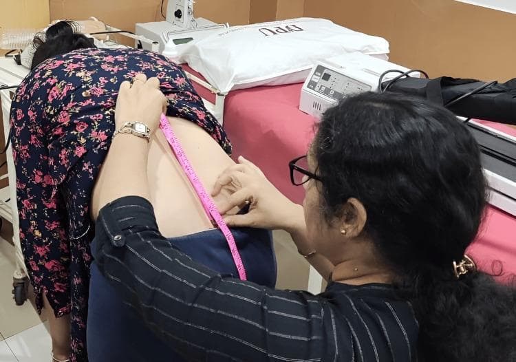This clinical photograph demonstrates the assessment of lumbar flexion range of motion using the Modified-Modified Schober’s Test (MMST). The patient is positioned in active forward trunk flexion, while the clinician, standing to the side and slightly behind, uses a pink flexible measuring tape to determine the distance between two pre-marked anatomical landmarks on the lumbar spine. These landmarks typically include a point level with the inferior margins of the posterior superior iliac spines (PSIS) and a second point marked 15 cm superiorly along the midline. The image captures the dynamic measurement phase where the expansion of the skin over the lumbar vertebrae is quantified. The setting is a physiotherapy clinic, indicated by a treatment table with a red cover, a pillow, and therapeutic modalities like a pressure biofeedback unit or electrical stimulation machine in the background. This procedure is a standard clinical method for objectively evaluating spinal mobility and monitoring progress in patients with low back pain or movement control impairments.