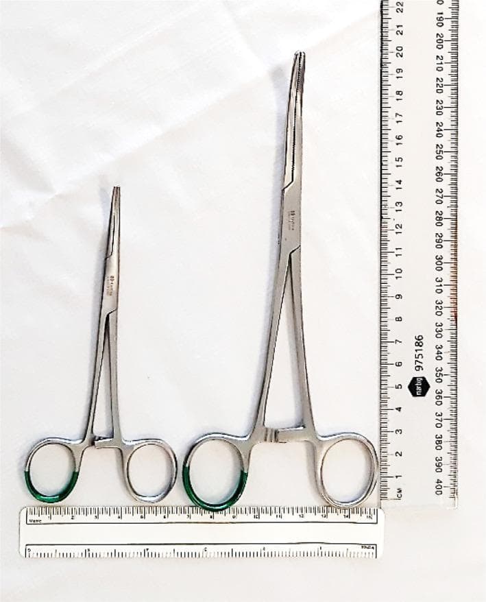 A top-down clinical photograph providing a side-by-side comparison of two common surgical hemostatic instruments: fine artery forceps (left) and Kelly clamps (right). Both stainless steel instruments feature finger rings with green identifiers, a ratchet locking mechanism, and longitudinal serrated jaws. The fine artery forceps on the left measure approximately 13.5 cm in total length with a delicate, tapered tip design. In contrast, the Kelly clamps on the right are significantly larger, measuring approximately 20 cm in length with broader, more robust jaws and a blunter tip. Rulers placed horizontally and vertically facilitate a scale comparison of the dimensions. This visual is intended to demonstrate the physical differences in instrument size and tip surface area, which are critical factors in surgical procedures such as parietal pleural puncture for chest tube insertion, where smaller instruments allow for more precise control and reduced force during tissue penetration.
