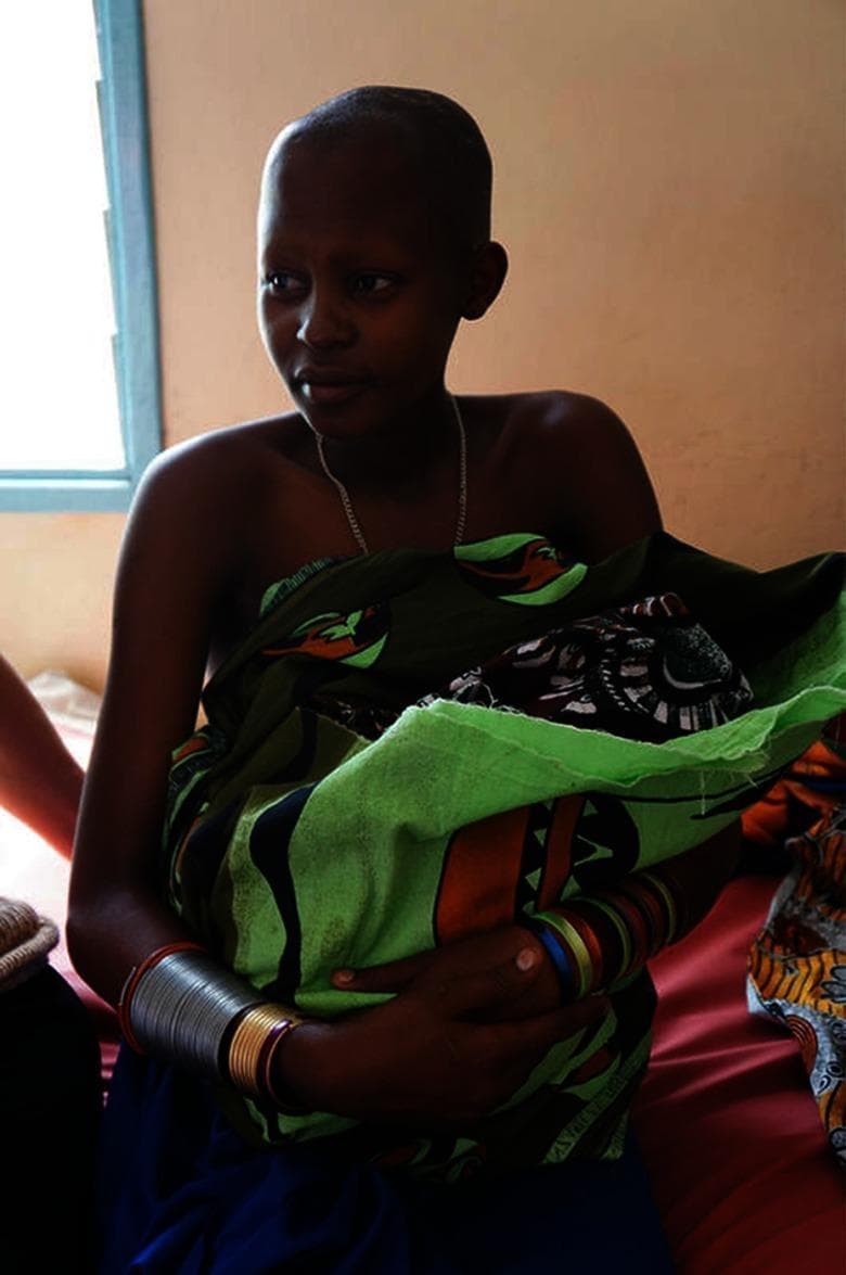 A clinical photograph depicting an adolescent postpartum patient in a district health facility. The image shows a 16-year-old female holding a neonate wrapped in a green and black patterned cloth. The patient is seated on a clinical examination bed or recovery cot covered with red linen, set within a simple medical room featuring a blue-framed window and peach-colored walls. The photograph serves as an educational visual for global maternal and child health, specifically highlighting the topics of adolescent pregnancy, postpartum care, and family planning in low-resource settings. Key educational concepts include the clinical management of young mothers and the social determinants of birth spacing as outlined in global health initiatives like Healthy People 2020. The setting represents a primary care or maternity ward context relevant to maternal-fetal medicine and reproductive health education.