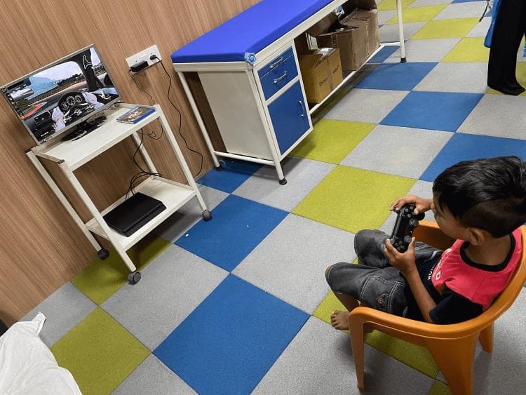 A clinical photograph illustrating a non-immersive virtual reality (NIVR) intervention setup for pediatric rehabilitation. The image depicts a child seated in a plastic chair, engaged with a driving simulation game using a handheld gaming controller. The rehabilitation apparatus consists of a mobile, two-tiered rolling cart. The upper shelf supports a digital monitor displaying the simulation and the lower shelf houses a PlayStation 4 console. The setting is a clinical therapy room, indicated by the presence of a professional medical examination table with a blue padded surface and storage cabinets. The intervention is utilized in physiotherapy to improve upper limb coordination, motor control, and functional independence in pediatric patients with conditions such as cerebral palsy. The floor features multi-colored tactile tiling, common in pediatric clinical environments.
