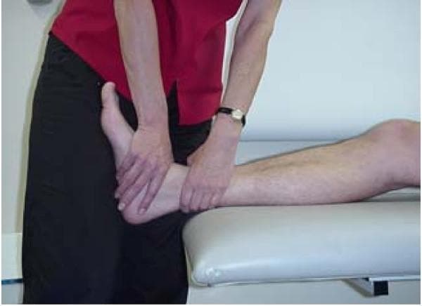 This procedural photograph illustrates a physiotherapist performing a passive joint mobilization technique on a patient's ankle, specifically an anterior-posterior (A-P) glide of the talus. The patient is positioned supine or in long-sitting on a treatment table with the lower limb extended. The clinician demonstrates a manual therapy technique where the left hand stabilizes the calcaneus and posterior ankle, while the right hand is positioned on the anterior aspect of the talus, just distal to the ankle joint line. This specific grip is used to apply Grade III oscillatory movements into resistance, aimed at improving dorsiflexion range of motion following injuries such as ankle sprains or fractures. The image serves as a clinical guide for orthopedic manual therapy, highlighting correct hand placement for mobilizing the talocrural joint.