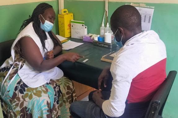 A clinical photograph depicting a post-test counseling session for a COVID-19 Rapid Diagnostic Test (RDT) in a community health setting. The image shows a healthcare worker or counselor and a client seated at an examination table, both wearing blue surgical face masks as personal protective equipment (PPE). The counselor points toward a lateral flow assay RDT cassette positioned on the table surface. The clinical workspace is organized with essential diagnostic supplies, including a yellow sharps disposal container for biohazardous waste, a box of test kits, bottles of hand sanitizer or reagent, swabs, and patient documentation on a clipboard. The image illustrates point-of-care testing protocols, infectious disease management, and the clinical communication process between health providers and patients regarding diagnostic results in a resource-limited setting.