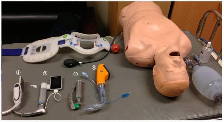 A clinical photograph of emergency airway management equipment and a training manikin for a comparative simulation study. The image displays four distinct laryngoscopes, numbered one through four, alongside adjunctive medical devices. Laryngoscope 1 is a McGrath MAC video laryngoscope (white body, integrated screen). Laryngoscope 2 is an AV-scope AVL (silver body with an attached rectangular monitor). Laryngoscope 3 is a conventional Macintosh laryngoscope (CML) with a metallic silver handle and a green-base blade. Laryngoscope 4 is a Pentax Airwayscope (orange handle with a transparent curved blade). Additional equipment includes a cervical collar with blue straps and color-coded adjustment indicators, a manual resuscitator bag (Ambu bag), and a medical training manikin torso. This setup is designed for clinical training and comparative evaluation of video laryngoscopy versus conventional direct laryngoscopy in scenarios involving airway stabilization. The educational focus is on intubation techniques, device morphology, and the selection of airway management tools for difficult airway scenarios or cervical spine immobilization training.