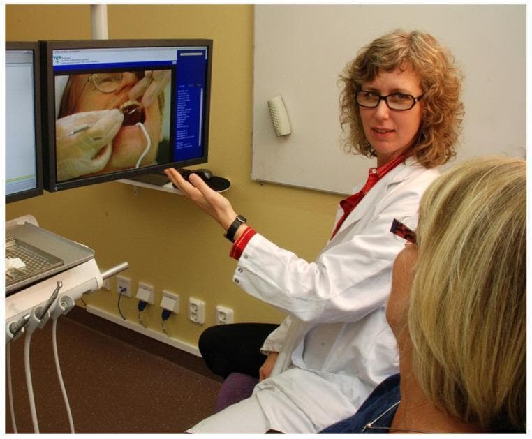 This clinical photograph illustrates a dental phobia treatment setting within a fully equipped dental operating room. A psychologist, identified by a white lab coat, is conducting an exposure-based Cognitive Behavioral Therapy (CBT) session for a patient with severe dental anxiety. The psychologist is gesturing toward a computer monitor that displays high-resolution imagery of a clinical dental procedure, serving as a visual exposure stimulus to desensitize the patient. The environment includes standard dental operatory equipment, such as an instrument console with handpieces and various dental tools, integrating the psychological intervention directly into the clinical space where dental triggers occur. This multidisciplinary approach aims to reduce avoidance behaviors and manage physiological anxiety reactions through gradual exposure and psychoeducation. The target audience for this material includes dental students, psychologists, and clinicians specializing in behavioral medicine or dental public health.