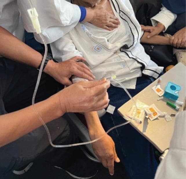 This clinical photograph captures a procedural scene of an intravenous (IV) cannulation and infusion being performed on a patient in a non-clinical environment, specifically an airplane cabin during a medical emergency. The image shows the insertion of an IV cannula into the patient's antecubital vein of the right arm, which is secured with white adhesive bandages. Clear plastic IV tubing with a visible drip chamber is connected to the cannula, facilitating the administration of normal saline and glucose. Two responders are visible: one in a white coat palpating the patient's upper chest/shoulder area, and another assisting by stabilizing the patient's arm and managing the infusion line. A small tray table in the foreground contains discarded medical supplies, including adhesive tape, alcohol swab wrappers, and a syringe. The photograph illustrates the clinical challenges of emergency medical intervention in confined spaces, emphasizing vascular access techniques for treating metabolic crises like diabetic emergencies in transit.