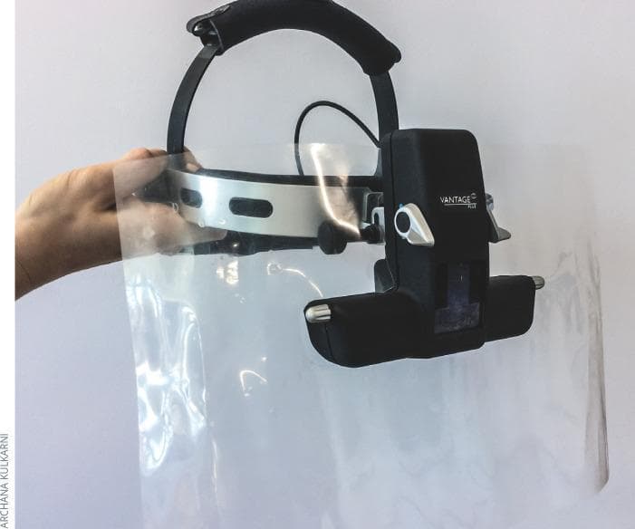 This clinical photograph displays a binocular indirect ophthalmoscope (BIO), specifically a Vantage Plus model, modified with a protective breath shield for infection control. The device features a black adjustable head strap with a padded crown and a silver metallic forehead band. The central optical unit is mounted to the front. Attached to this assembly is a large, clear, flexible plastic breath shield that extends downward and laterally to provide a physical barrier between the ophthalmologist and the patient. A hand is shown supporting the transparent shield to demonstrate its flexibility and wide coverage area. This modification is used in ophthalmology to minimize the risk of respiratory droplet transmission, particularly during close-proximity examinations required for retinal evaluation. The setup represents a standard preventative measure used in clinical settings to maintain hygiene and safety during diagnostic procedures.