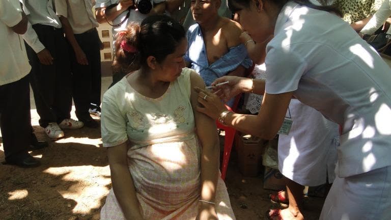 This clinical photograph depicts a public health immunization outreach scene in a rural setting in Vietnam. A seated pregnant woman is receiving an intramuscular vaccination, such as the influenza vaccine, administered by a health professional. The healthcare worker, dressed in a white clinical uniform with a visible identification tag, is performing the injection into the patient's deltoid muscle using a standard syringe and needle. The surrounding environment illustrates a community-based grassroots health system, featuring other residents waiting for care and an outdoor village backdrop. The image serves as an educational example of maternal immunization programs, primary healthcare delivery in resource-limited or mountainous regions, and the role of village health workers in promoting public health and preventive medicine during pregnancy.