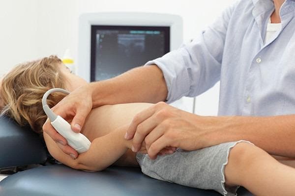 This clinical procedural photograph demonstrates the point-of-care ultrasound (POCUS) technique for diagnosing a 'pulled elbow' (radial head subluxation) in a pediatric patient. The child is positioned supine on a medical examination table with the head turned laterally. A medical professional is seen applying a high-frequency linear ultrasound transducer to the posterior-lateral aspect of the patient's elbow joint. The probe is oriented caudally to visualize the radiohumeral joint and the annular ligament. In the background, an ultrasound monitor displays a real-time grayscale musculoskeletal image. This procedure is used in pediatric emergency medicine to identify signs such as an increased distance between the radial head and the humerus capitate or the interposition of the annular ligament, which confirms subluxation. The image illustrates proper hand placement for stabilizing the young patient's limb while manipulating the transducer to achieve diagnostic views.