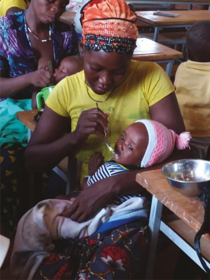 This clinical photograph illustrates a key aspect of pediatric nutrition and public health, depicting a woman feeding an infant in Burkina Faso. The image serves as an educational resource regarding the importance of complementary feeding and animal-sourced foods in low- and middle-income countries. The caregiver is using a metal spoon to feed the infant from a metal bowl containing eggs, which are high-quality sources of protein, essential amino acids, and polyunsaturated fatty acids like docosahexaenoic acid (DHA) and eicosapentaenoic acid (EPA). These nutrients are critical for neurodevelopment and preventing growth stunting. The setting appears to be a communal or educational environment, highlighting maternal-child health interventions. Key educational concepts include dietary diversity, the role of micronutrients (Vitamin A, B12, iron, and zinc) in preventing protein-energy malnutrition and anemia, and the impact of animal-sourced foods on cognitive development and overall physical growth during the first 1,000 days of life.