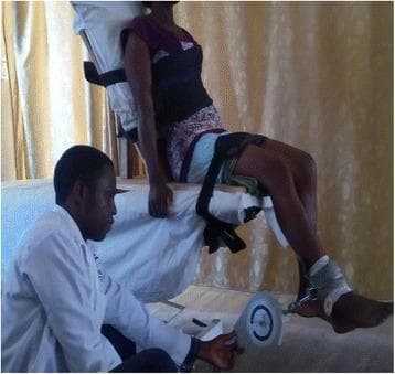 A clinical photograph depicting a participant undergoing static quadriceps muscle strength testing in a clinical or research setting. The individual is seated upright on a testing table with the back firmly supported against a vertical backrest. Safety and stabilization straps are visible across the chest and thighs to prevent compensatory movements. The participant's knees are positioned at the edge of the table, flexed at approximately 60 degrees. A cable tensiometer is attached to the participant's distal lower leg using a padded ankle cuff. This device measures isometric force (kg or Newtons) as the participant attempts to extend the knee against the resistance of the fixed cable. A clinician in a white lab coat is shown monitoring the tensiometer scale. This setup is typical for physical therapy assessments and rehabilitation research to evaluate quadriceps peak force and muscle function, particularly in the management of knee osteoarthritis or post-surgical recovery.