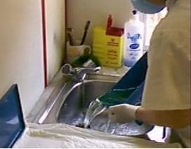 A clinical photograph depicting the maintenance and disinfection protocol in a healthcare setting. A healthcare worker, wearing appropriate personal protective equipment (PPE) including a face mask, white clinical coat, and surgical gloves, is seen rinsing a blue glutaraldehyde soaking tray in a stainless steel laboratory sink. The person is emptying residual high-level disinfectant solution under a running faucet. The background illustrates a controlled medical environment containing a yellow sharps disposal container for biohazardous waste, bottles of chemical disinfectants, and cleaning supplies. This image illustrates occupational safety practices and the procedural steps for the manual decontamination and cleaning of medical equipment used in sterilization or high-level disinfection processes. Key educational concepts include chemical safety, PPE compliance, and medical device reprocessing workflows.