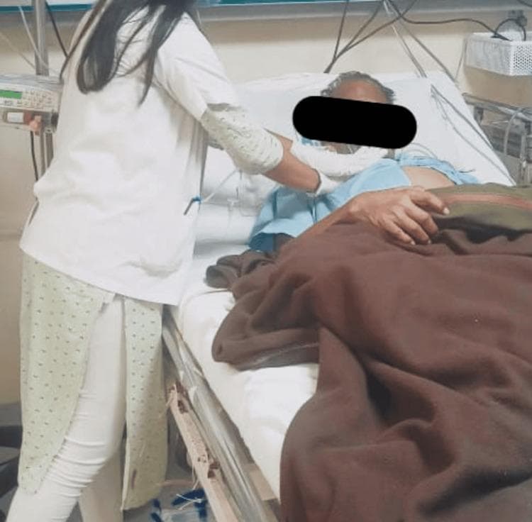 This clinical photograph illustrates a healthcare professional, likely a physical therapist, performing chest physiotherapy on a patient in an inpatient setting. The clinician is seen standing at the bedside, applying manual chest vibration techniques to the patient's anterior thoracic region. The patient is positioned supine in a hospital bed, equipped with side rails and integrated medical devices. Visible equipment includes a bedside monitor or infusion pump on a vertical pole, along with various wires and tubing consistent with continuous clinical monitoring or oxygen therapy. The educational focus of the image is on respiratory rehabilitation interventions, specifically the manual clearance of secretions (airway clearance therapy) in patients with conditions like pneumonia or chronic obstructive pulmonary disease. The image demonstrates the practical application of chest PT, emphasizing the tactile and manual nature of vibration and percussion to facilitate mucus drainage and improve pulmonary function.